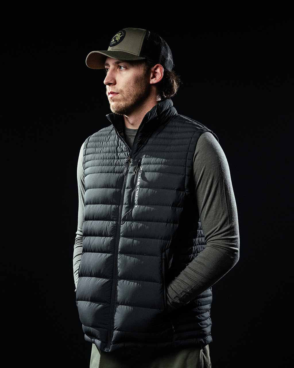 Ein Mann in einer EBERLESTOCK McCall Down Vest - Black, die über einem langärmeligen Hemd liegt und eine Baseballmütze trägt, steht mit den Händen in den Taschen und blickt zur Seite vor einem schwarzen Hintergrund.