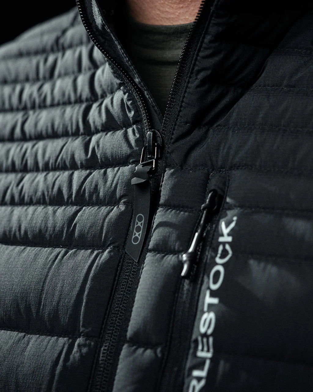 Nahaufnahme einer Person, die die EBERLESTOCK McCall Down Vest - Black trägt, wobei der Reißverschluss teilweise geöffnet ist. Die Weste ist mit grauen Gänsedaunen mit 850er Füllung ausgestattet und das EBERLESTOCK-Logo ist auf der rechten Seite teilweise in Weiß zu sehen.