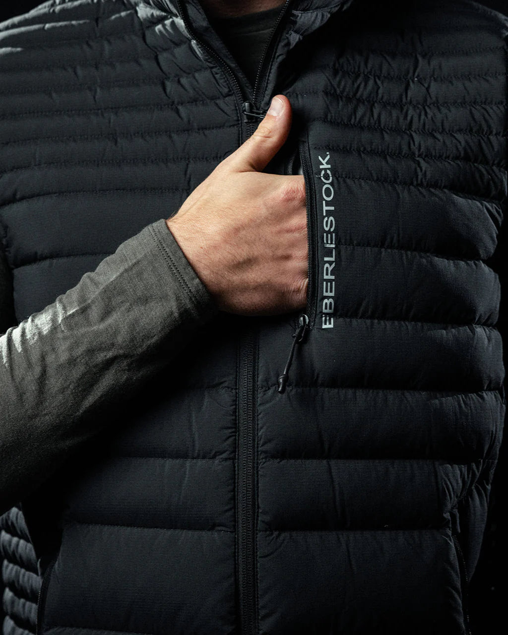 Eine Person in einer schwarzen EBERLESTOCK McCall Down Vest öffnet den Reißverschluss mit einer Hand und gibt den Blick frei auf das vertikale Markenlogo auf der Brusttasche und die 850er Gänsedaunen für hervorragende Wärme.