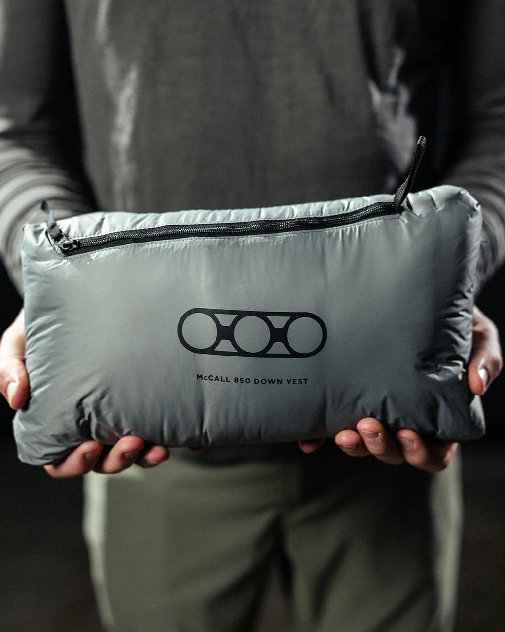 Eine Person hält eine verpackte schwarze EBERLESTOCK McCall Daunenweste in einer Reißverschlusstasche mit Markenlogo, die mit einem langärmeligen Hemd und einer grünen Hose gestylt ist. Die Premium-Weste ist mit 850er Daunen gefüllt und wärmt.