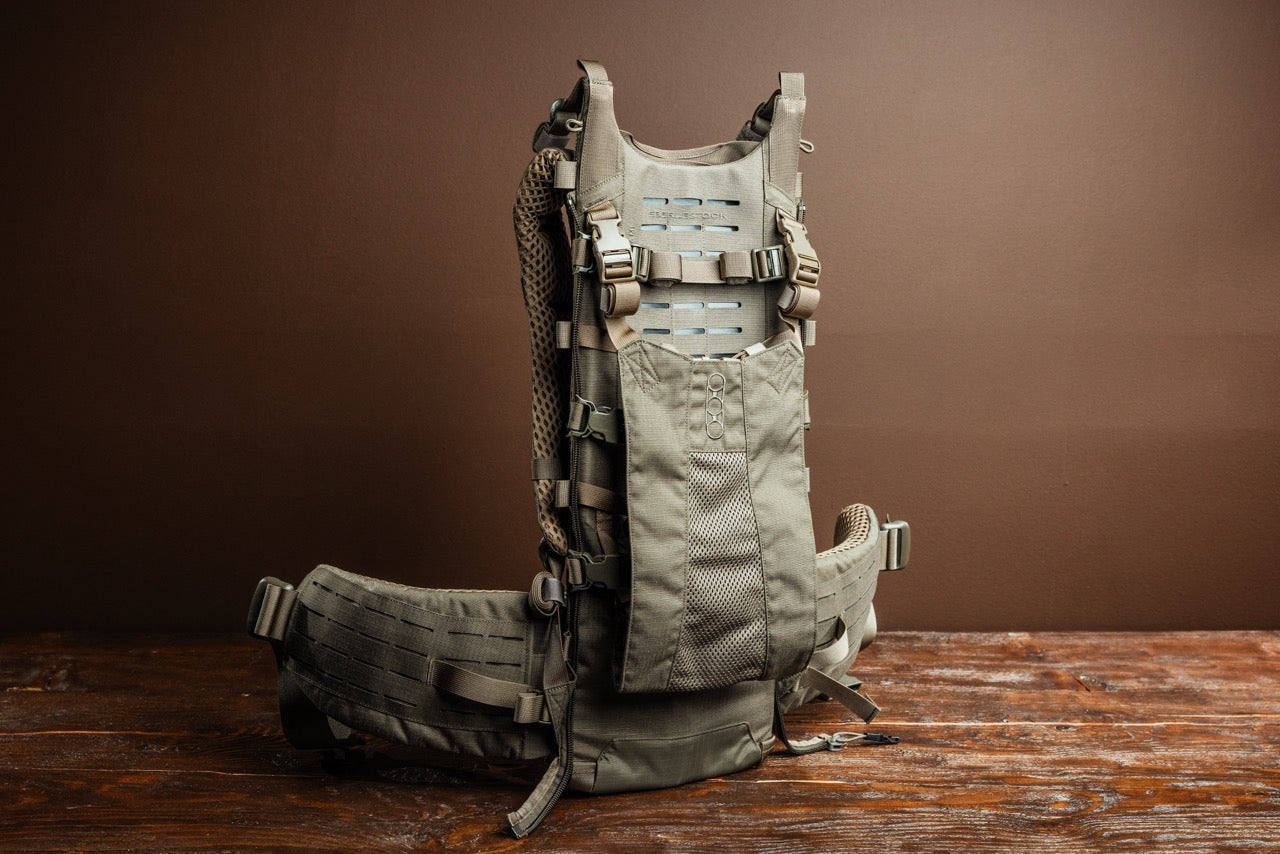 Der EBERLESTOCK Modframe - Military Green Rucksack mit mehreren Riemen, Schnallen und Netzpolsterung steht aufrecht auf einer Holzoberfläche mit braunem Hintergrund. Dieser modulare Rucksack ist auf Langlebigkeit und Outdoor-Einsatz ausgelegt.