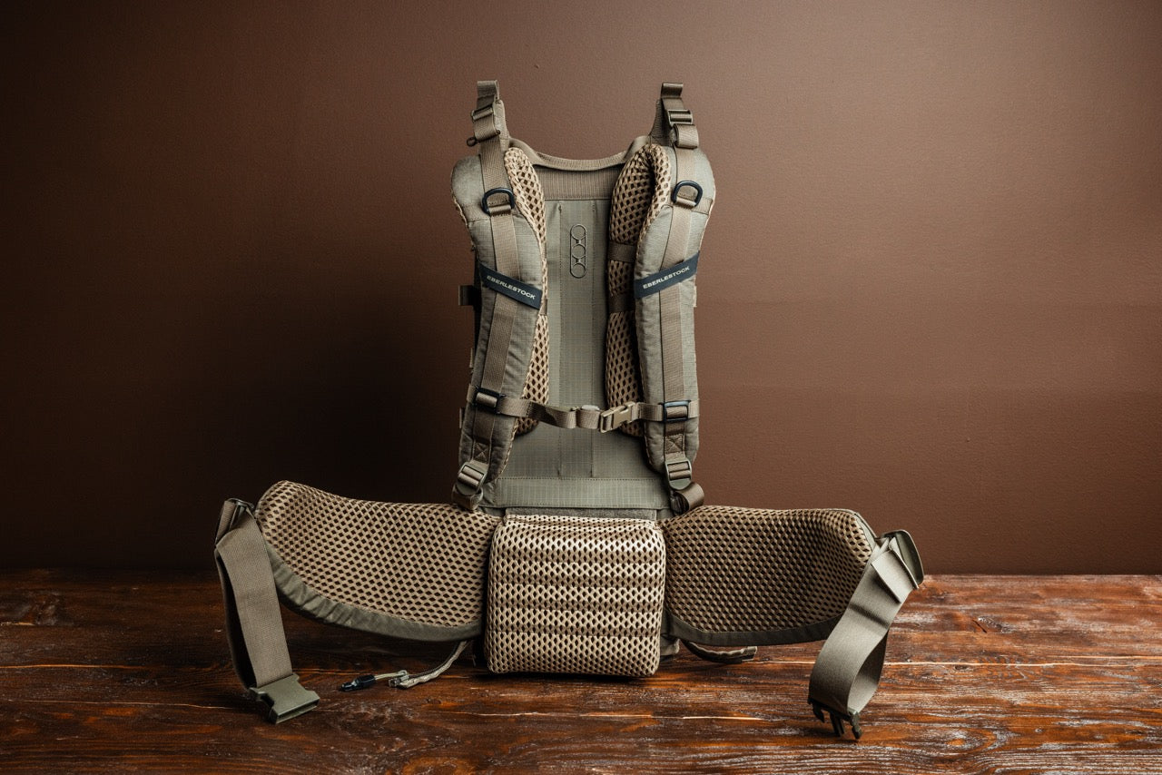 Der EBERLESTOCK Modframe in Military Green ist aufrecht auf einer Holzoberfläche mit braunem Hintergrund abgebildet. Er verfügt über Mesh-Polsterung, verstellbare Schultergurte und einen breiten gepolsterten Hüftgurt für Komfort und Halt.