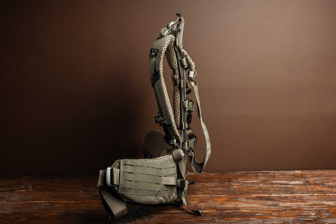 Der EBERLESTOCK Modframe - Military Green Rucksackrahmen verfügt über Mesh-Polsterung, verstellbare Gurte, Schnallen und einen gepolsterten Hüftgurt. Er steht aufrecht auf einer hölzernen Oberfläche vor einem braunen Hintergrund.