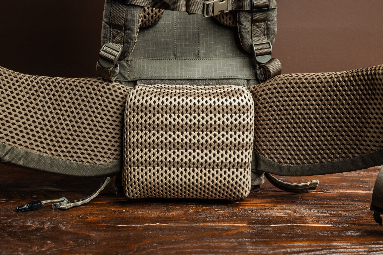 Nahaufnahme des gepolsterten Mesh-Hüftgurts und -Rückenteils des EBERLESTOCK Modframe - Military Green mit atmungsaktivem, strukturiertem Material und stabilen Riemen auf einer Holzoberfläche mit braunem Hintergrund.