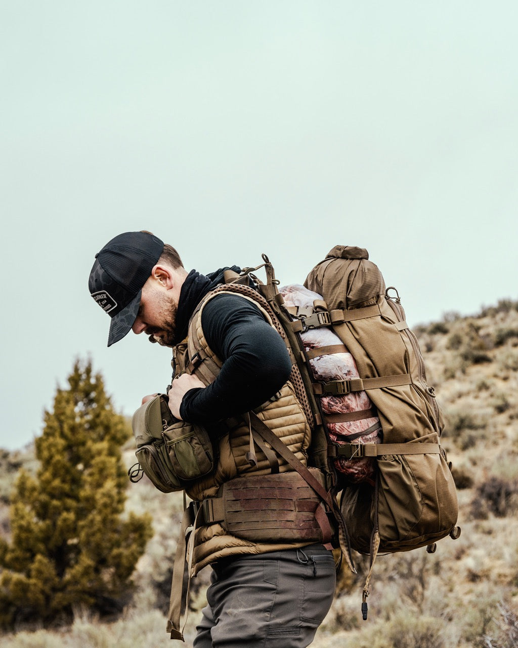 Ein Mann in Outdoor-Ausrüstung und mit schwarzer Mütze trägt einen EBERLESTOCK Modframe - Military Green Rucksack und eine kleinere Tasche in einer rauen, grasbewachsenen Landschaft mit Büschen und einem kleinen Baum im Hintergrund.