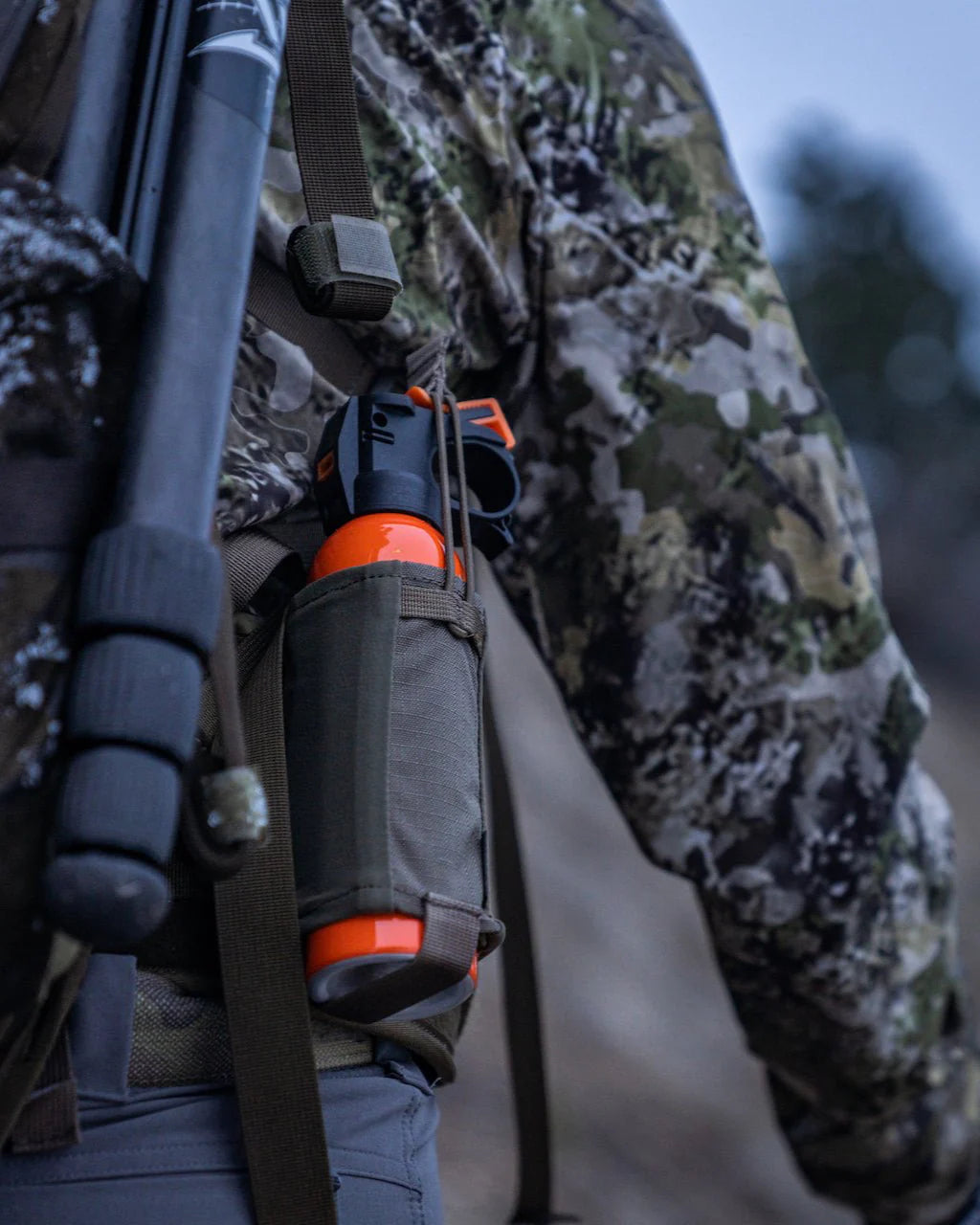 Eine Person in Tarnkleidung trägt eine Schrotflinte und ein Bärenspray in einer EBERLESTOCK Recon Bear Spray Pouch, die am Rucksackgurt befestigt ist, während sie im Freien unterwegs ist.