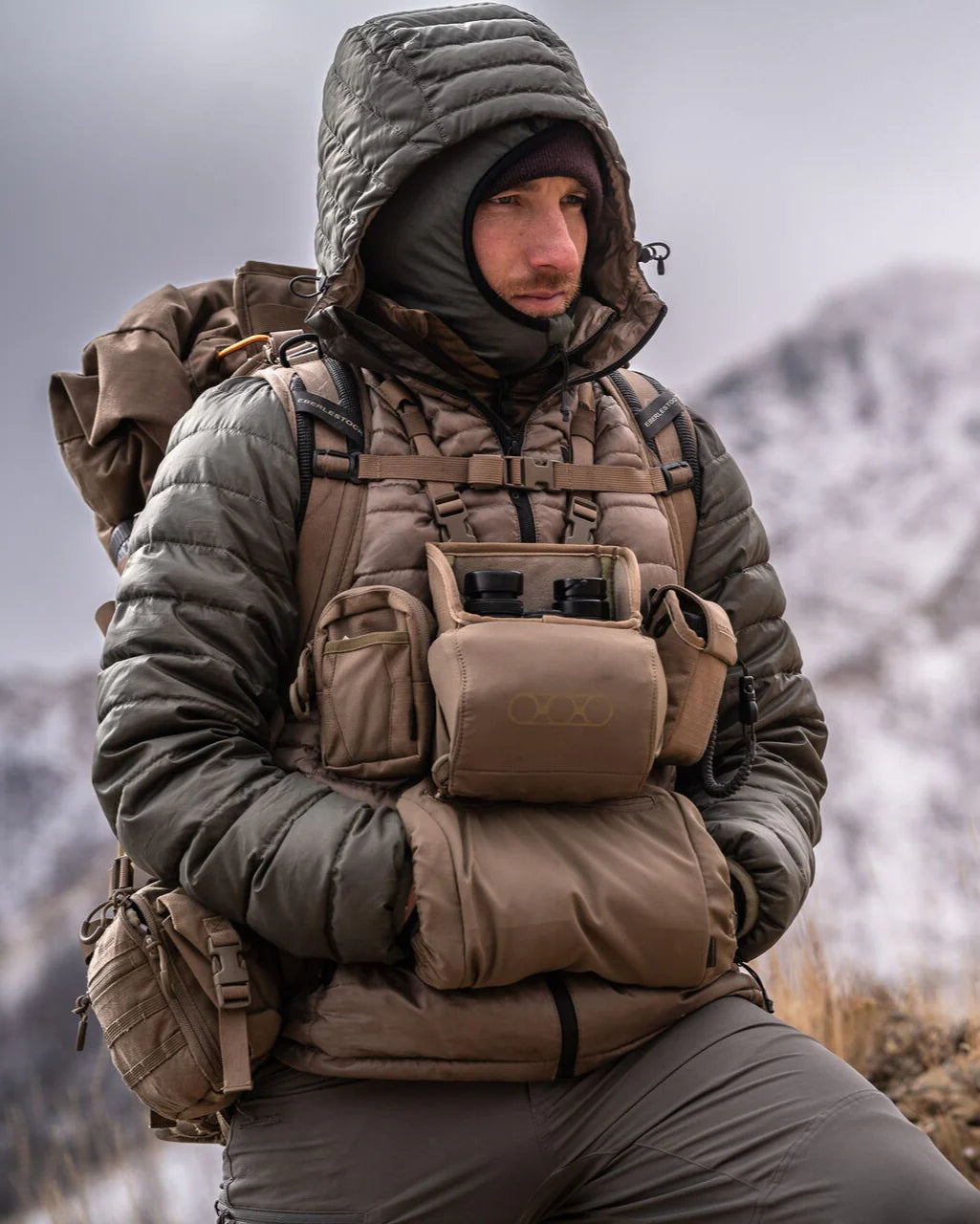 Eine Person mit Kapuzenpulli-Jacke steht in den verschneiten Bergen, trägt einen großen Rucksack und benutzt den EBERLESTOCK Recon Handwarmer, um die Hände beim Wandern in der Kälte warm zu halten.