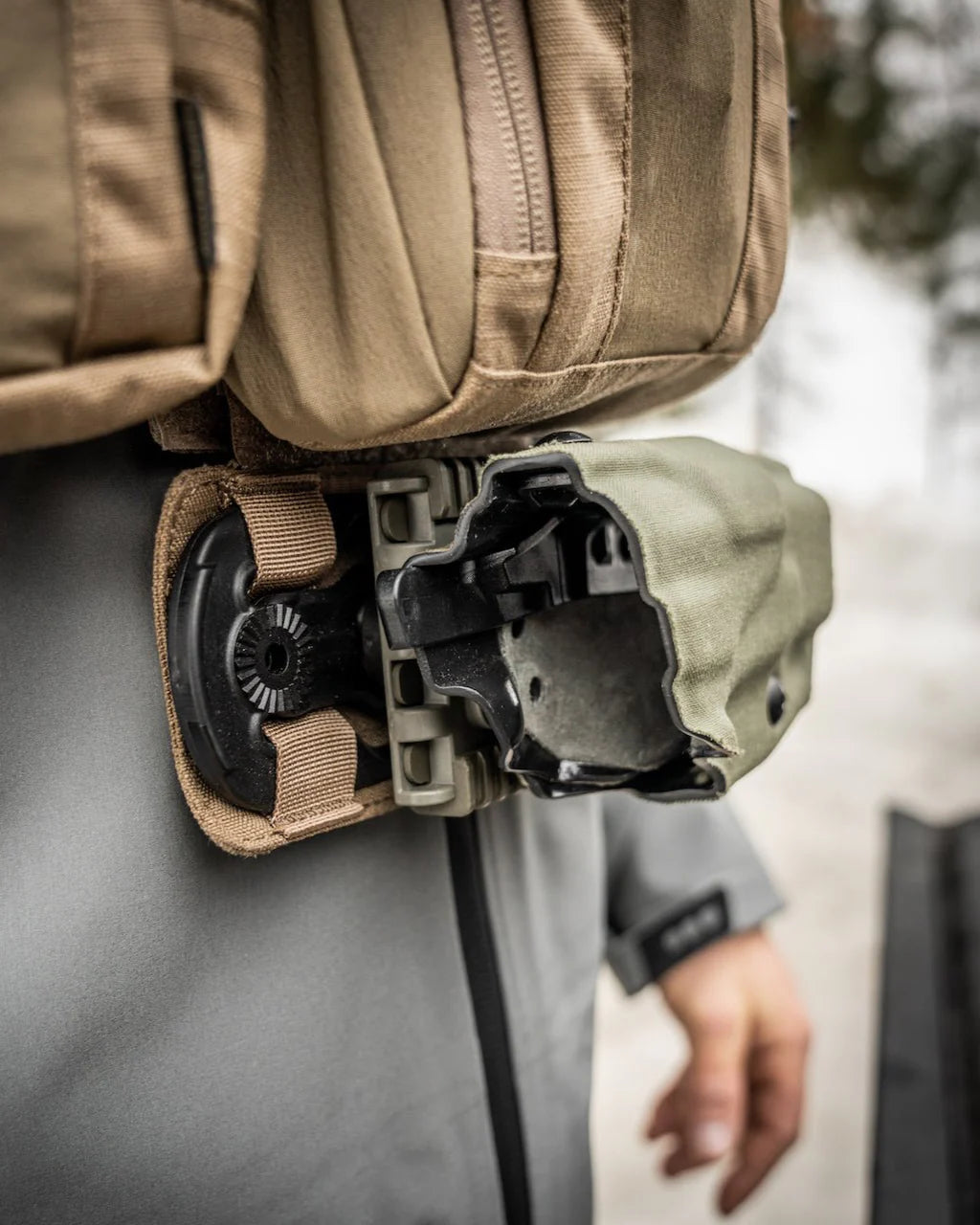 Nahaufnahme einer Person in taktischer Ausrüstung mit einem hellbraunen und grünen Hartschalenholster an der Hüfte neben einem EBERLESTOCK Recon MOLLE Panel, die Hand teilweise im Hintergrund sichtbar.