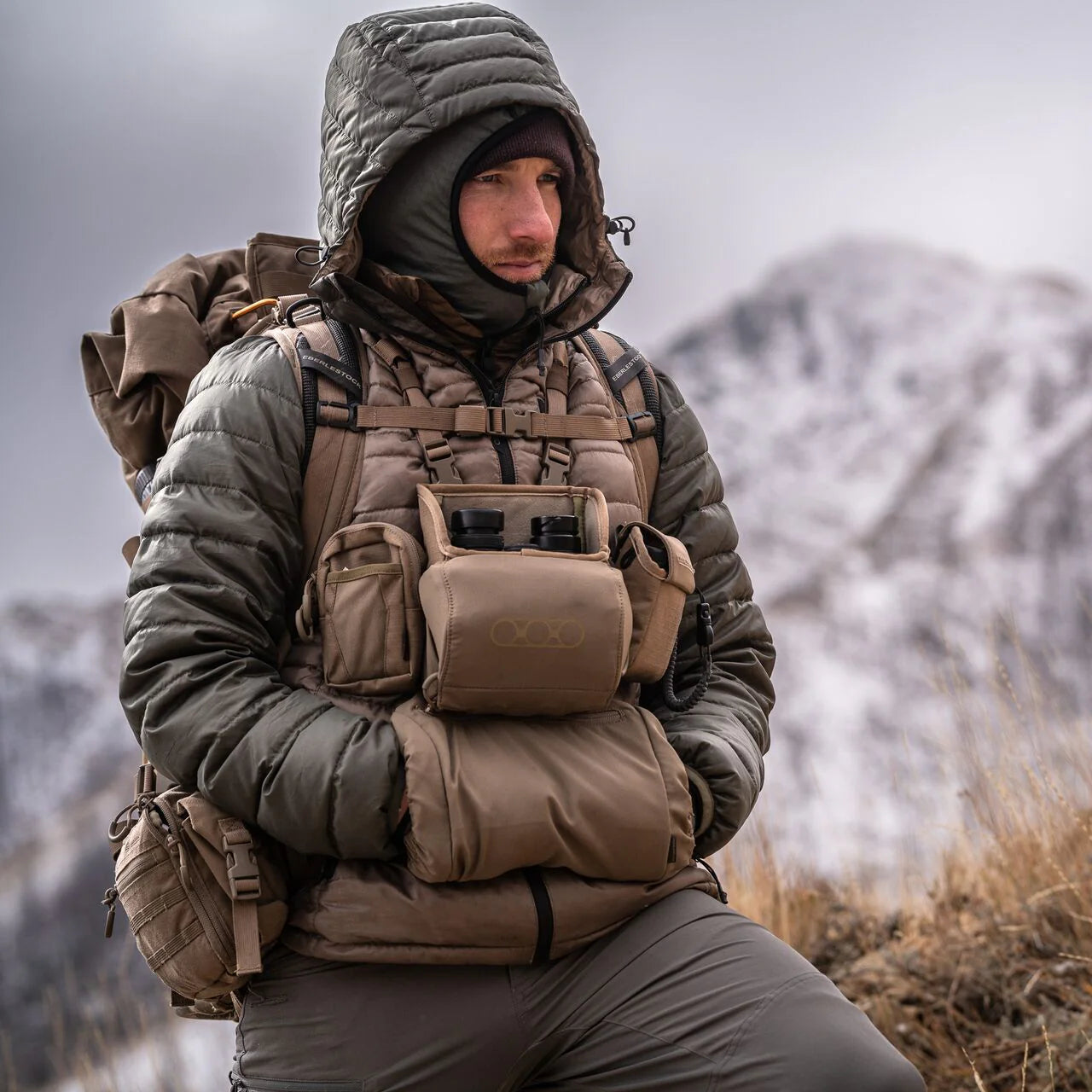 Bekleidet mit isolierter Outdoor-Ausrüstung und Handschuhen steht eine Person mit einem großen Rucksack und dem EBERLESTOCK Recon Modular Bino Pack Large in einer verschneiten Berglandschaft und blickt in die Ferne.