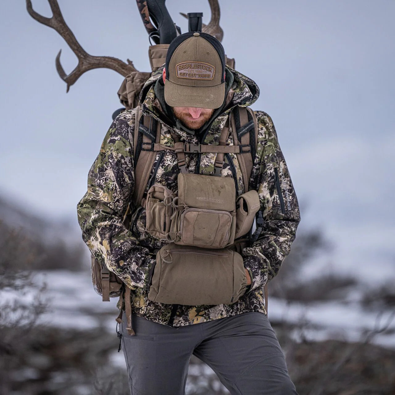 In Tarnfarben und mit einer Mütze bekleidet steht eine Person in einer verschneiten, zerklüfteten Landschaft mit einem großen Rucksack, der mit einem Geweih verziert ist. Sie trägt das EBERLESTOCK Recon Modular Bino Pack Small - eine ideale Wahl für jedes Jäger Outdoor Abenteuer.