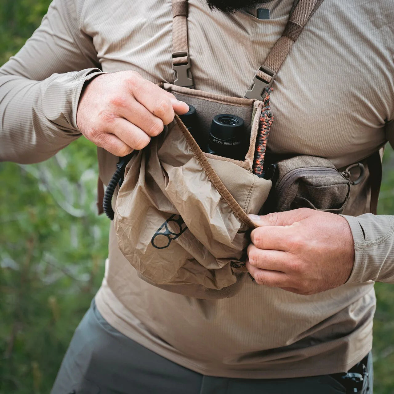 Eine Person in einem hellbraunen, langärmeligen Hemd öffnet eine braune Tasche, die an einer EBERLESTOCK Recon Rain Fly befestigt ist, und gibt den Blick auf eine Brille und ein schwarzes, zylindrisches Objekt frei, mit verschwommenem Grün im Hintergrund.
