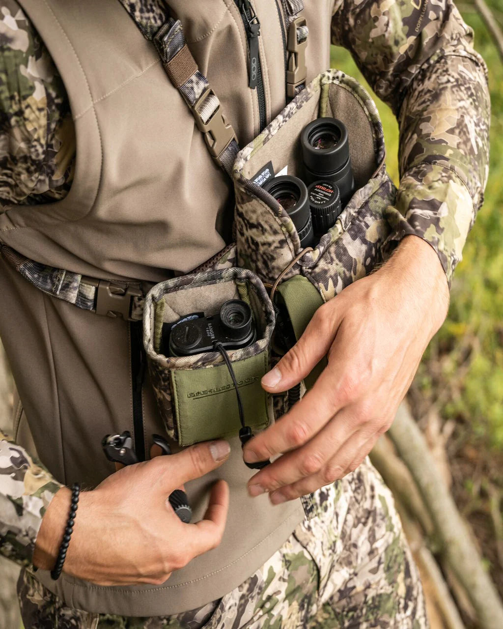 Eine Person in Tarnkleidung hält im Freien inmitten von grünem Laub ein Fernglas in der Hand, das teilweise in einer EBERLESTOCK Recon Rangefinder Pouch an ihrer Weste verstaut ist.