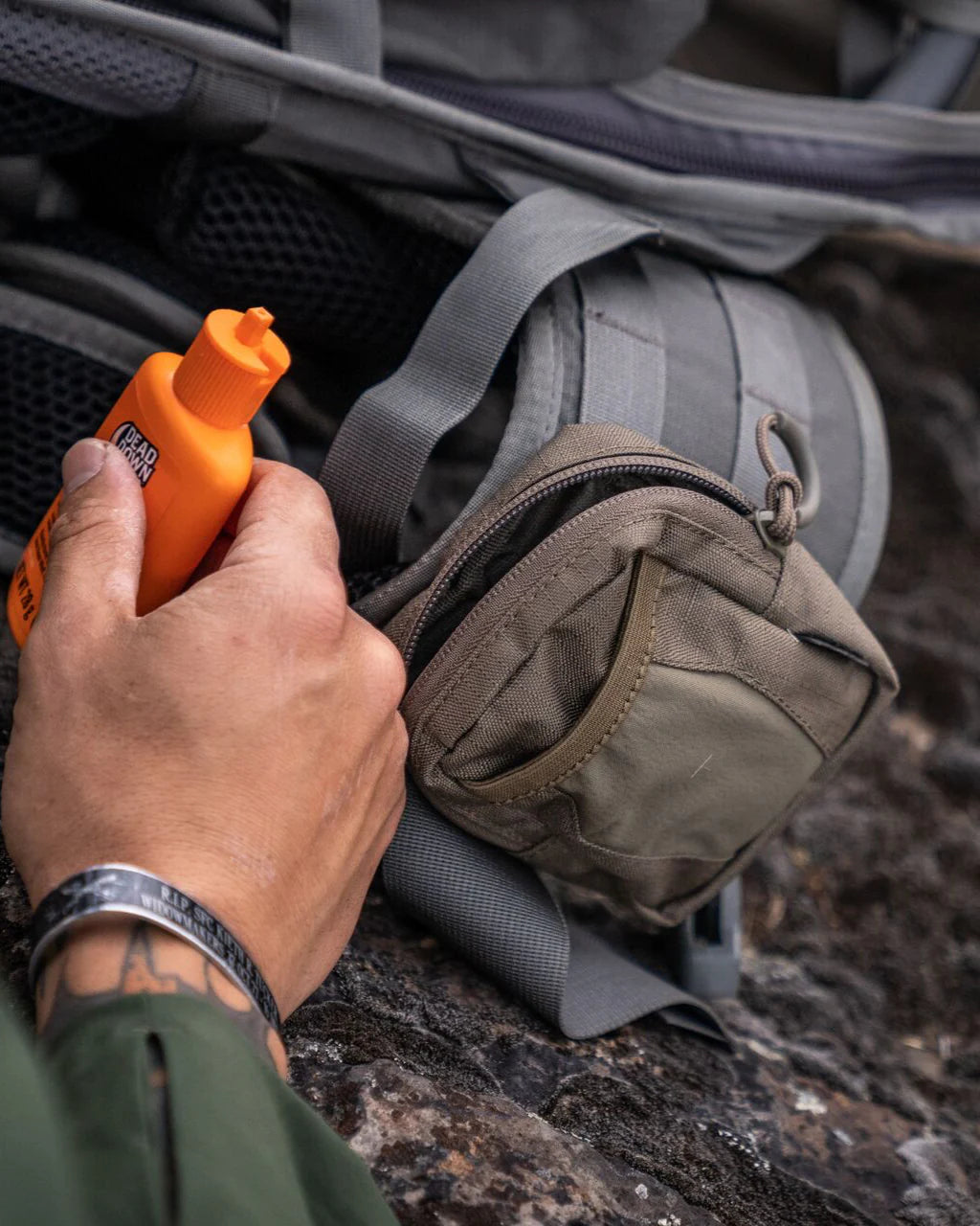 Eine Person hält eine orangefarbene Flasche mit Insektenschutzmittel neben einer kleinen EBERLESTOCK Recon Utility Pouch, während sie im Freien sitzt und sich auf eine Wanderung vorbereitet.