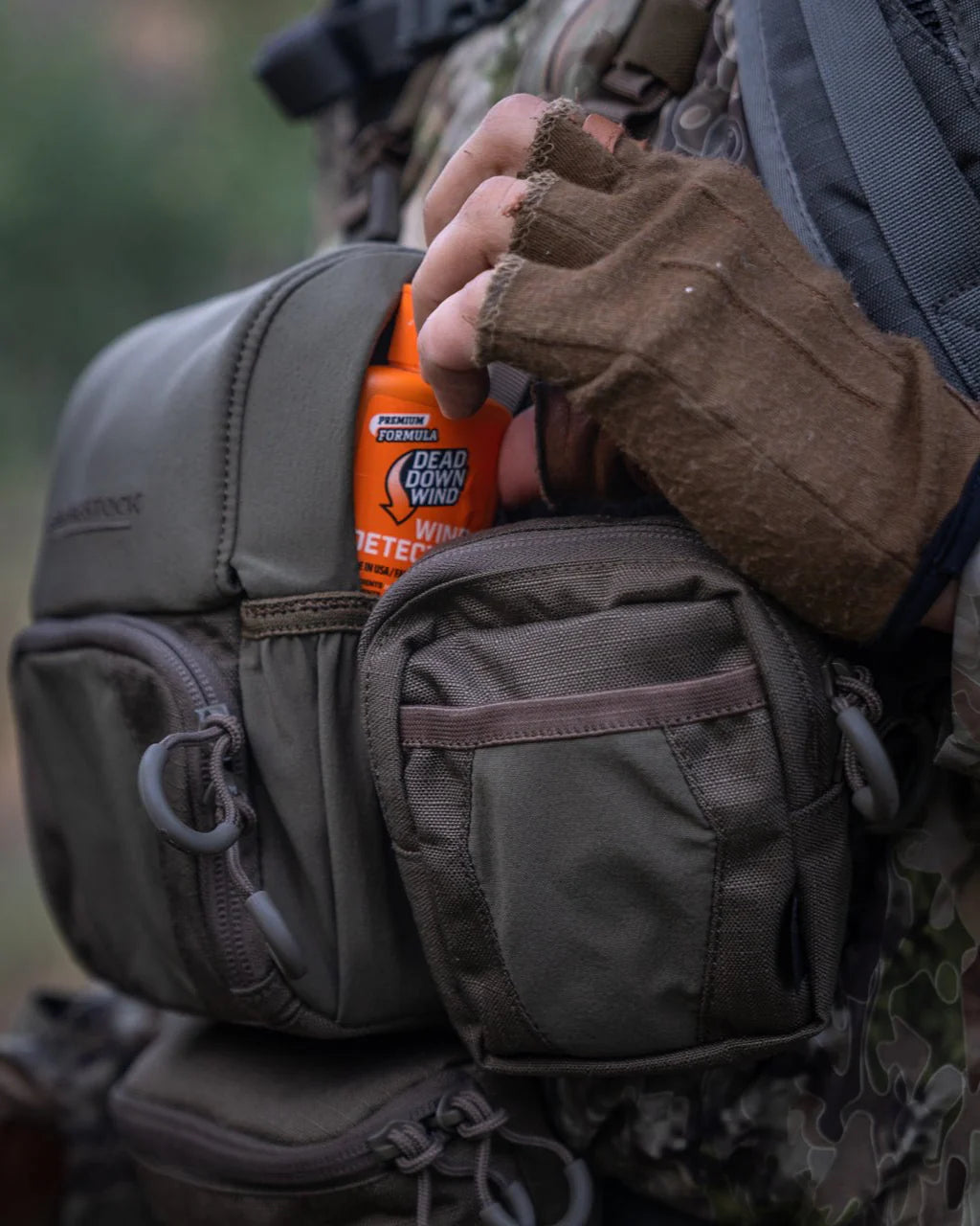 Eine Person in Tarnkleidung und fingerlosen Handschuhen steckt eine Flasche Geruchseliminierer in eine EBERLESTOCK Recon Utility Pouch, die an ihrer MOLLE-kompatiblen taktischen Weste befestigt ist.