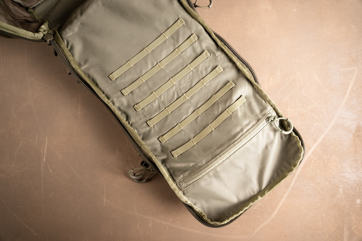 Ein offener schwarzer EBERLESTOCK Romad Rucksack liegt flach und offenbart sein Inneres mit mehreren horizontalen MOLLE-Gurten und Fächern zur Organisation auf einer hellbraunen Oberfläche.