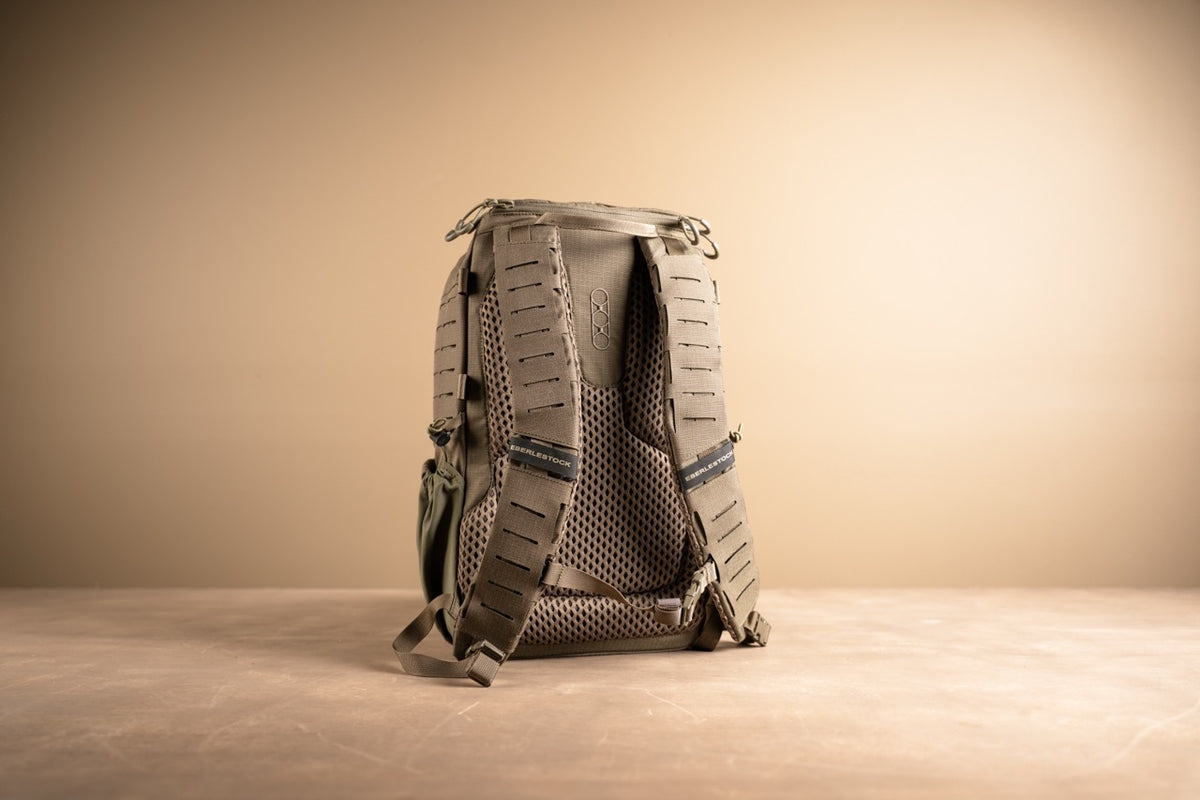 Der EBERLESTOCK Romad Rucksack in Schwarz steht aufrecht auf einer neutralen Oberfläche vor einem beigen Hintergrund und zeigt seinen ergonomischen Rücken, gepolsterte Mesh-Gurte, mehrere Schlaufen und MOLLE-Panel.
