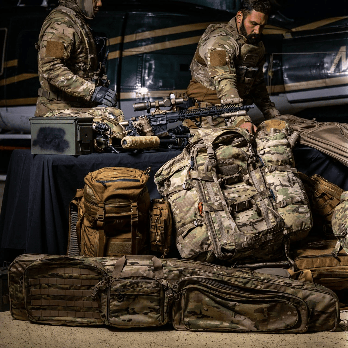 Zwei Personen in militärischer Tarnkleidung stehen an einem Tisch mit taktischen Rucksäcken, einem EBERLESTOCK Sniper Sled Drag Bag 52", einem Gewehr und Ausrüstungskoffern, während im Hintergrund ein Hubschrauber zu sehen ist.