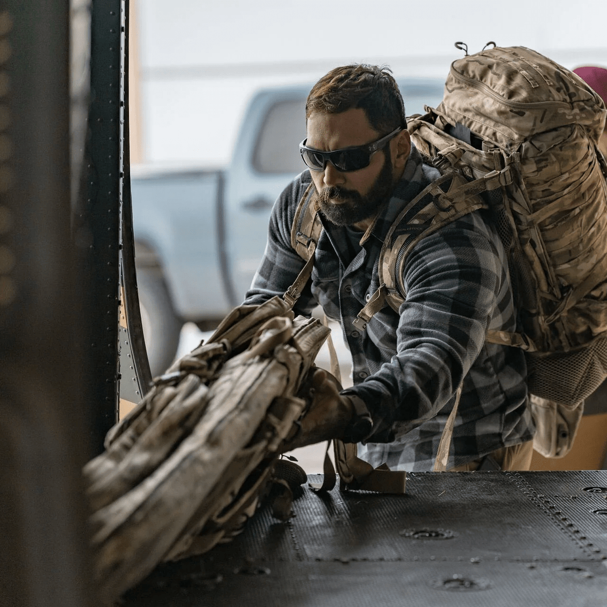 Ein Mann mit Sonnenbrille, kariertem Hemd und großem Rucksack lädt seine Ausrüstung - darunter die EBERLESTOCK Sniper Sled Drag Bag 52" - auf ein Fahrzeug oder Flugzeug, während im Hintergrund ein blauer Lastwagen zu sehen ist.