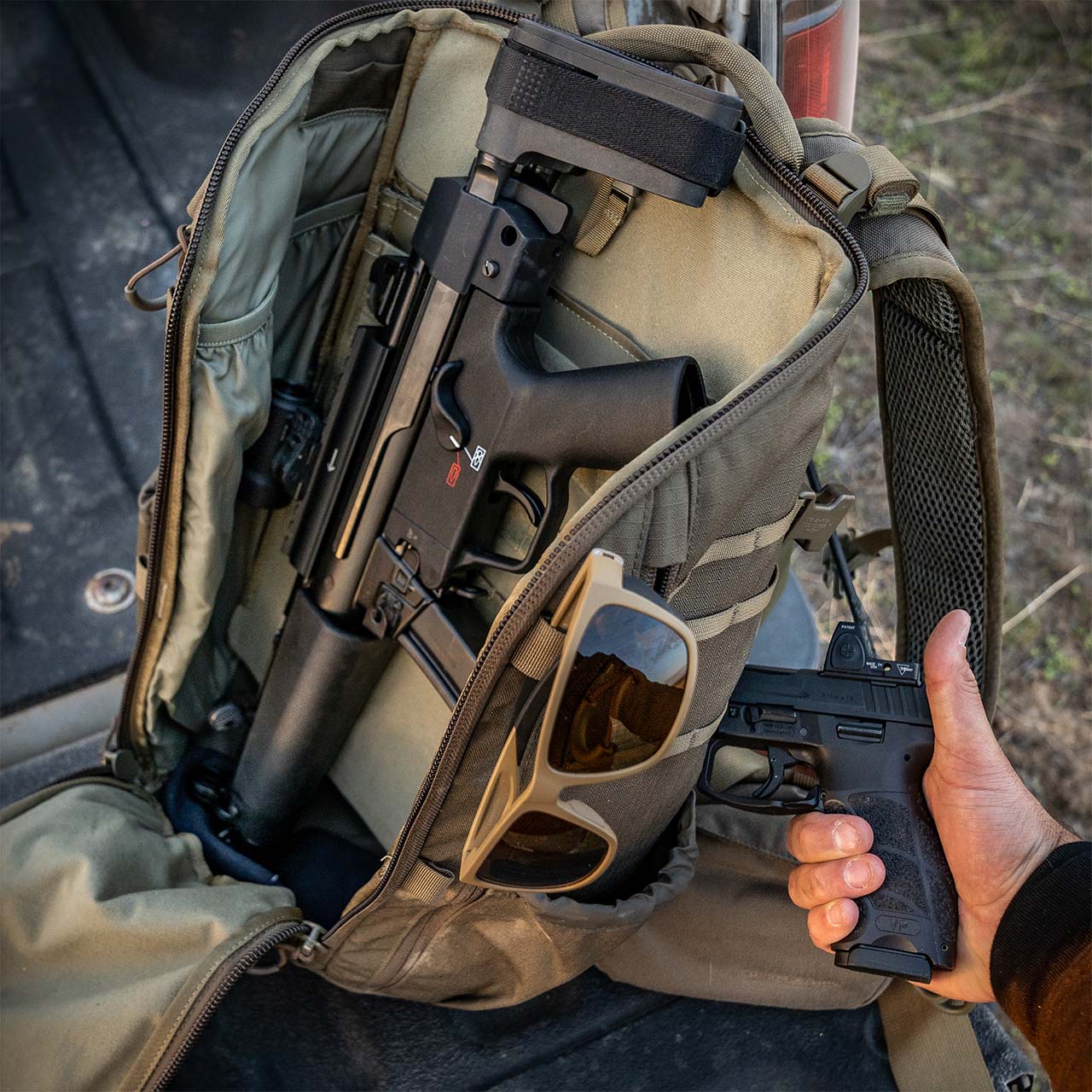 Ein kompakter Rucksack, wie der EBERLESTOCK Eberlestock Switchblade Pack aus 500D-Nylon, ist geöffnet und zeigt darin eine schwarze Maschinenpistole. In der Nähe hält eine Hand eine schwarze Pistole, Sonnenbrillen liegen auf der Fronttasche.