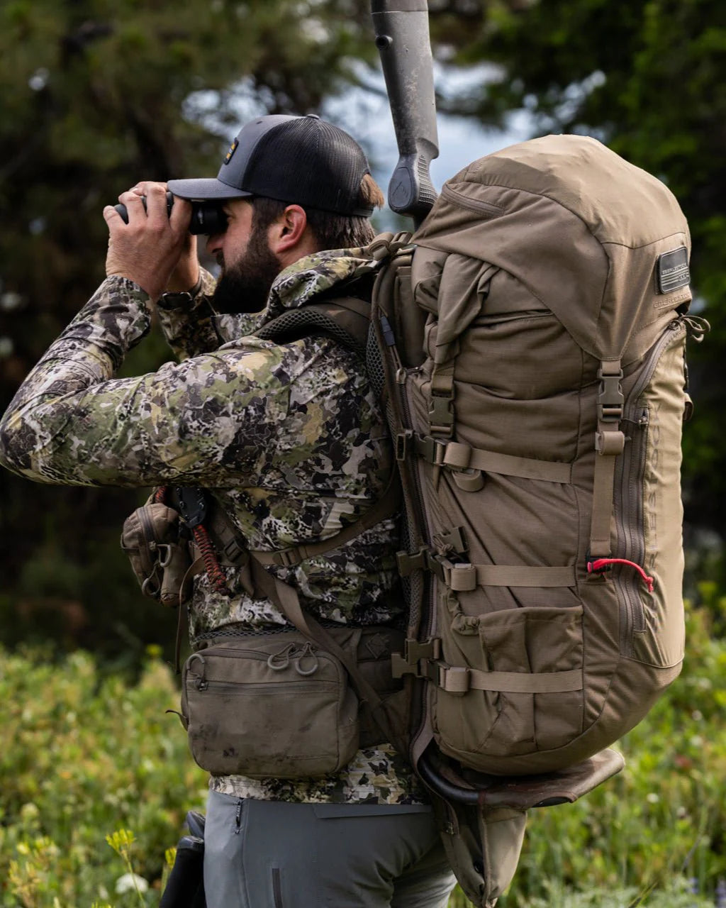 Eine Person in Camouflage und mit schwarzer Mütze schaut durch ein Fernglas ins Freie und trägt den EBERLESTOCK Vapor 5000 V2 (82L) Rucksack mit Ausrüstung, umgeben von grünen Bäumen und Gras.