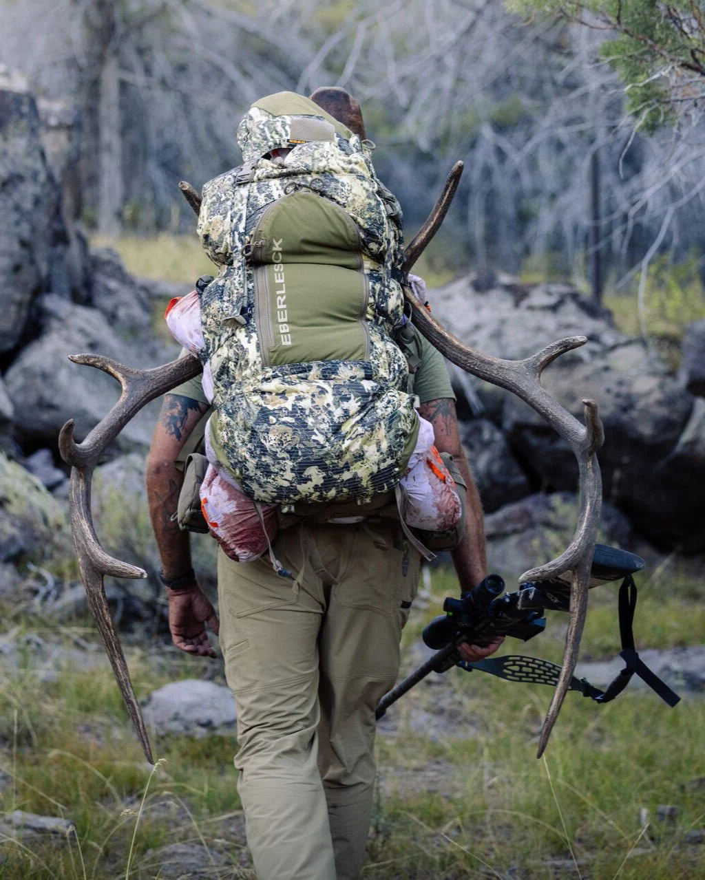 Eine Person in Tarnkleidung trägt den EBERLESTOCK Vapor 5000 V2 Rucksack (82 l) mit Geweih und eingewickeltem Wildfleisch, während sie durch felsiges Gelände läuft und einen Bogen hält.
