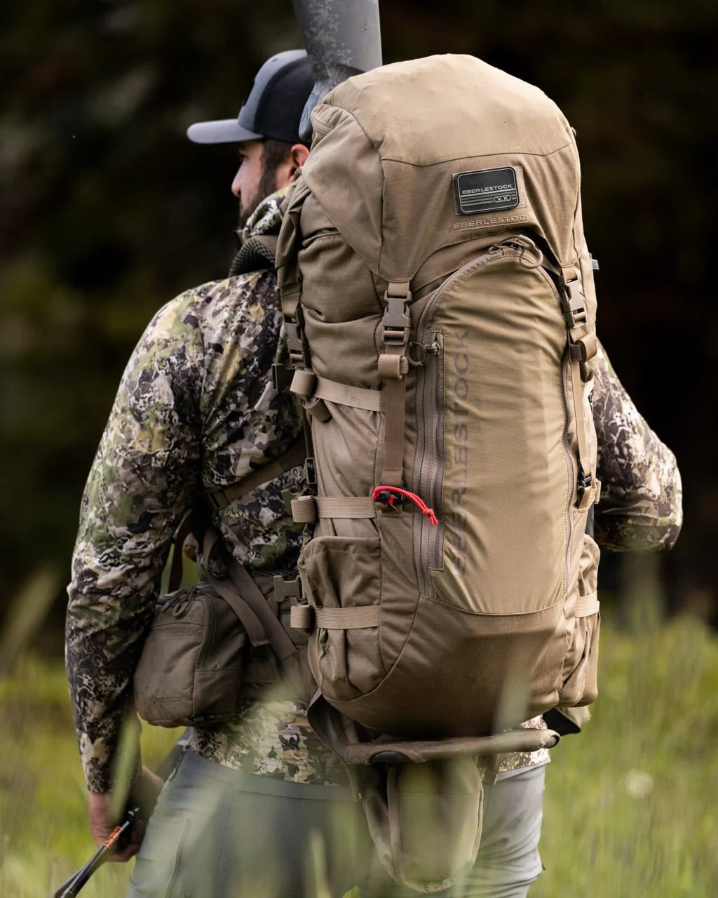 Eine Person in Camouflage und mit Mütze steht im Freien auf einer Wiese und trägt den EBERLESTOCK Vapor 5000 V2, einen ultraleichten 82-Liter-Rucksack mit mehreren Riemen, einem Markenaufnäher und einem rot akzentuierten Reißverschlusszug.