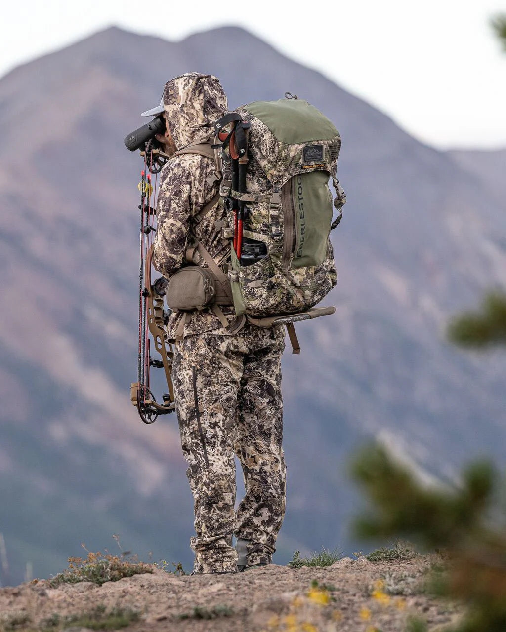 In voller Tarnkleidung steht eine Person im Freien mit einem EBERLESTOCK Vapor 5000 V2 Rucksack (82L), hält einen Bogen und benutzt ein Fernglas, mit unscharfen Bergen im Hintergrund.