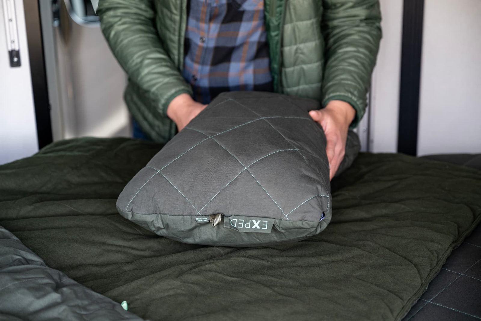 Eine Person in grüner Jacke legt das graue, gesteppte EXPED Kissen LuxeWool Pillow aus Bio-Baumwolle auf einen dunkelgrünen Schlafsack in einem Wohnmobil.