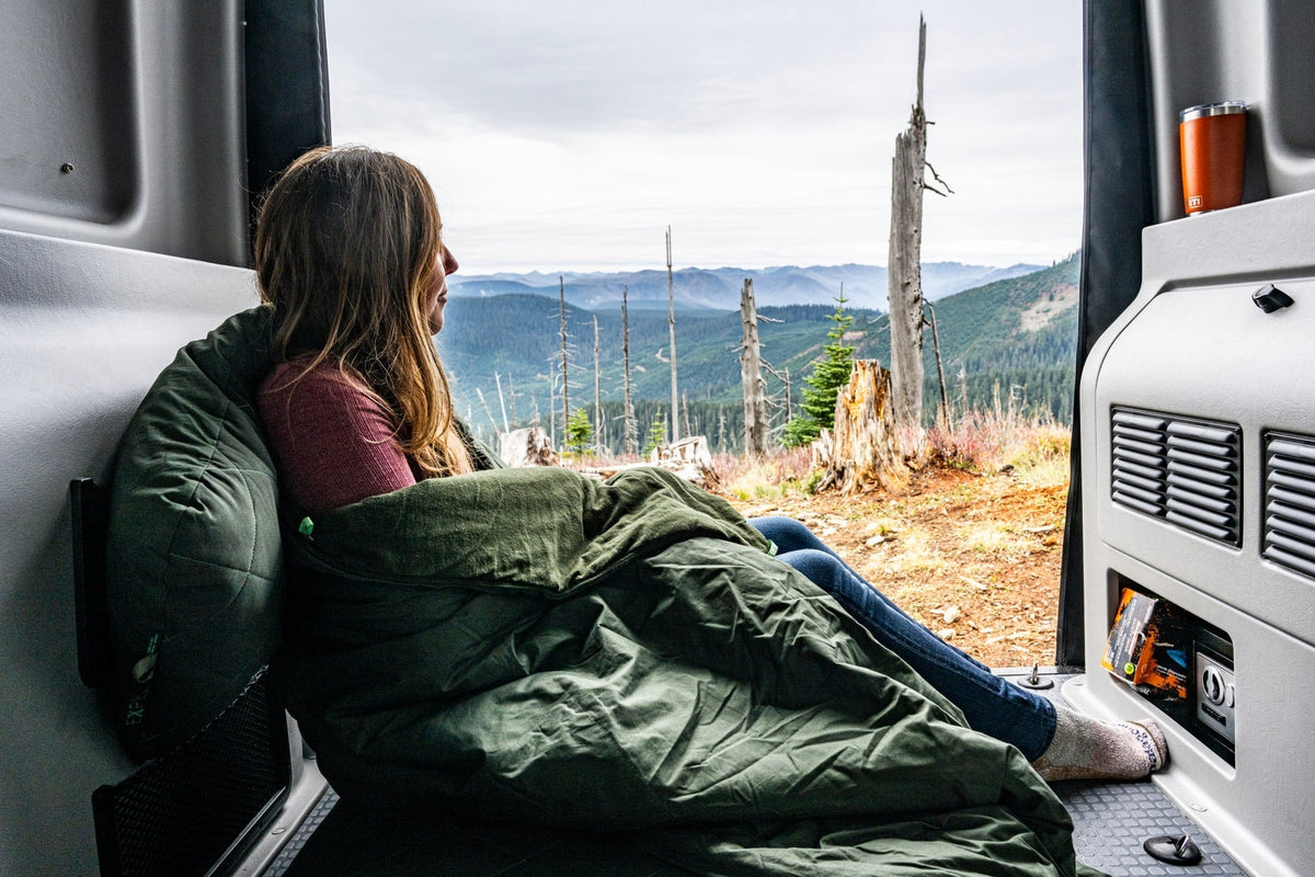 Eine Frau in einem grünen Schlafsack entspannt sich im Inneren eines Lieferwagens und ruht sich auf einem EXPED Kissen LuxeWool Pillow aus, während sie durch die offenen Hintertüren unter einem wolkenverhangenen Himmel auf die malerischen Berge und Bäume blickt.