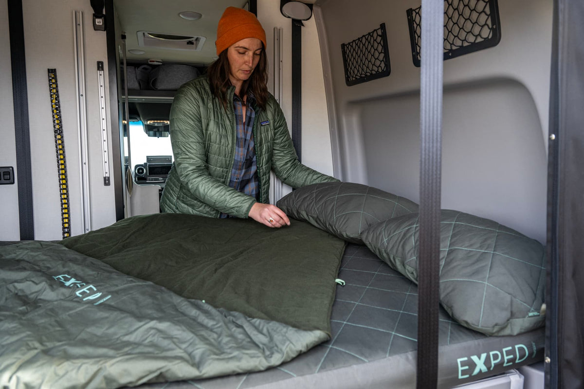 Eine Person in grüner Jacke und orangefarbener Mütze macht ein Bett in einem Wohnmobil und legt grüne Bio-Baumwolle-Bettwäsche und ein EXPED Kissen LuxeWool Pillow auf die Matratze. Dahinter sind Stauräume und das Innere des Wohnmobils zu sehen.