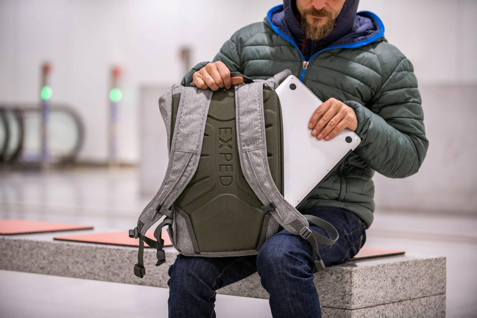 Ein Mann in einer grünen Pufferjacke sitzt auf einer Bank in einem modernen Innenraum und packt ein weißes Tablet oder einen Laptop in seinen grau-grünen EXPED Rucksack Metro 30.
