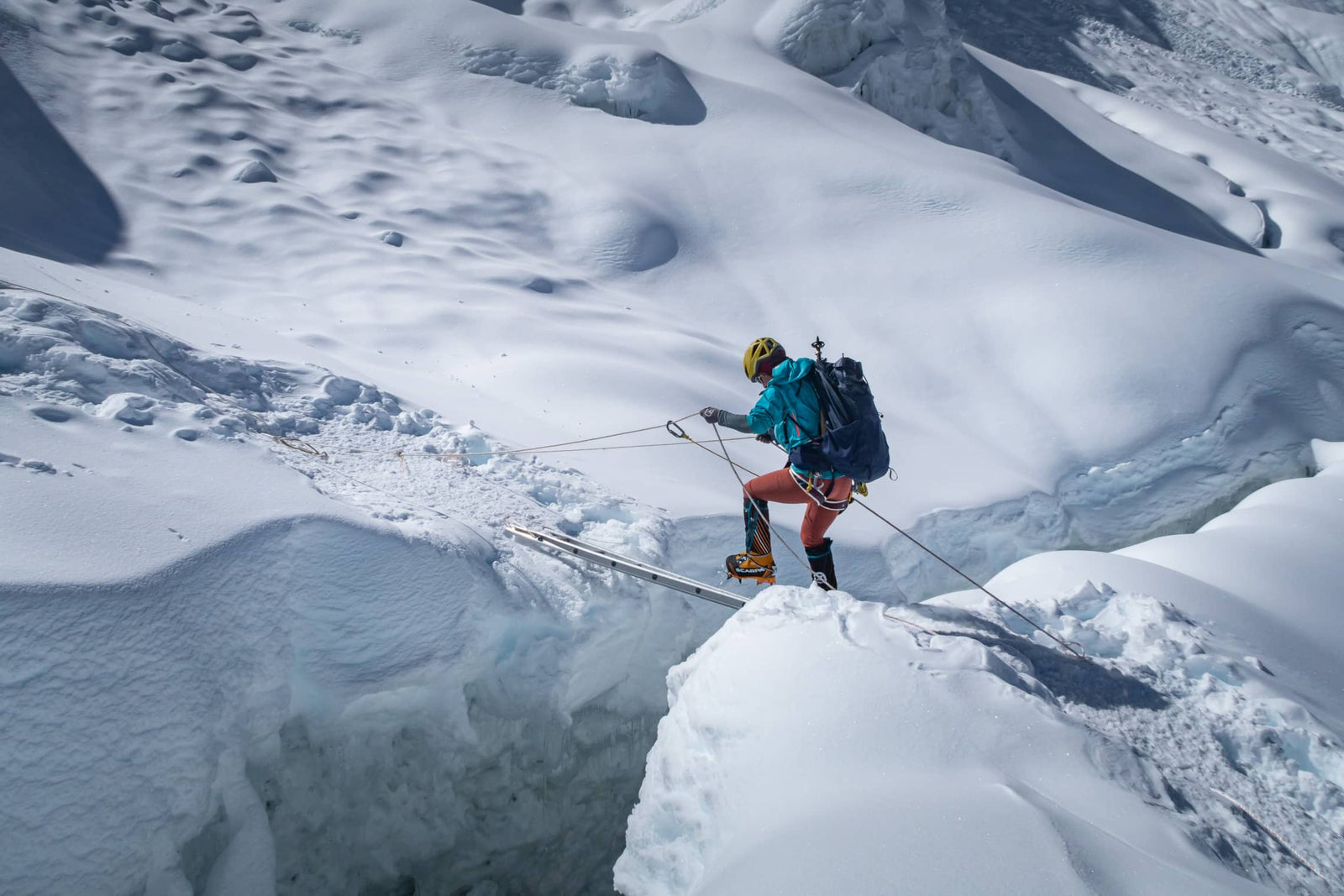 Ein Bergsteiger mit einem EXPED Rucksack Thunder 50 und Helm überquert mit Hilfe einer Leiter und Seilen vorsichtig eine tiefe Gletscherspalte auf einem verschneiten Berg. Das eisige Gelände deutet auf anspruchsvolle Mehrtagestouren hin.