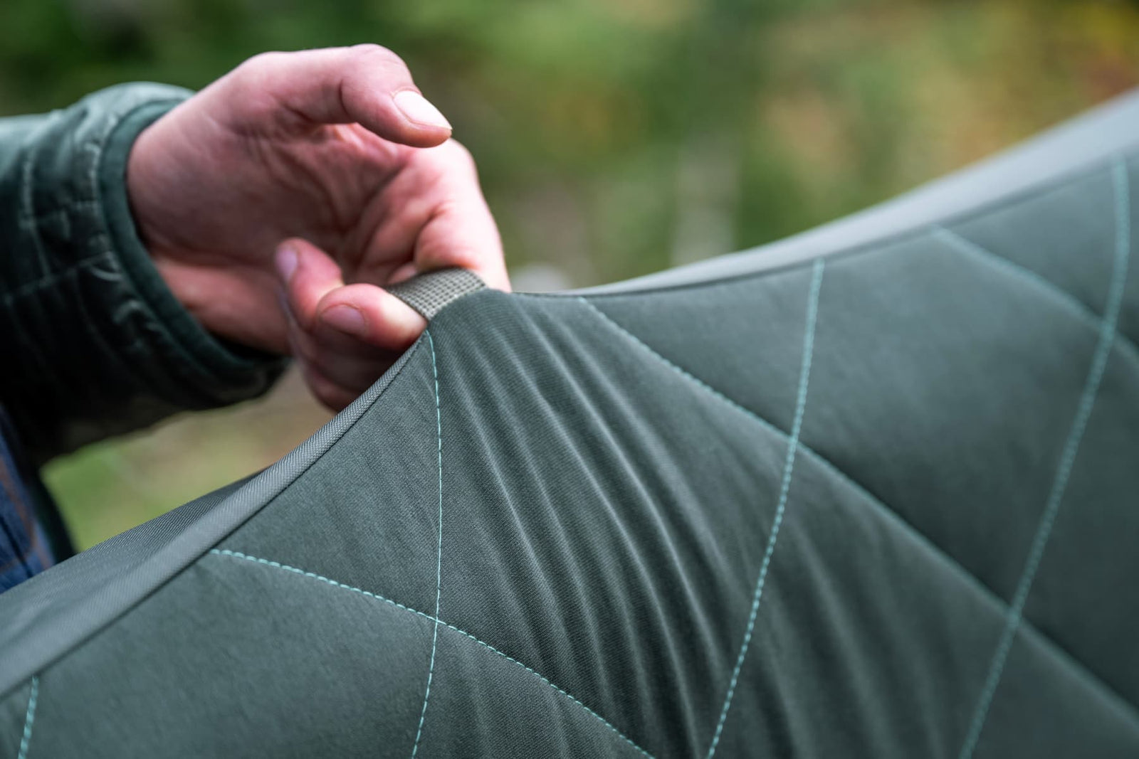 Eine Hand greift in die Ecke einer EXPED Schlafmatte LuxeMat DUO LW+, einer gesteppten grau-grünen Matte, mit verschwommenem grünem Laub im Hintergrund.