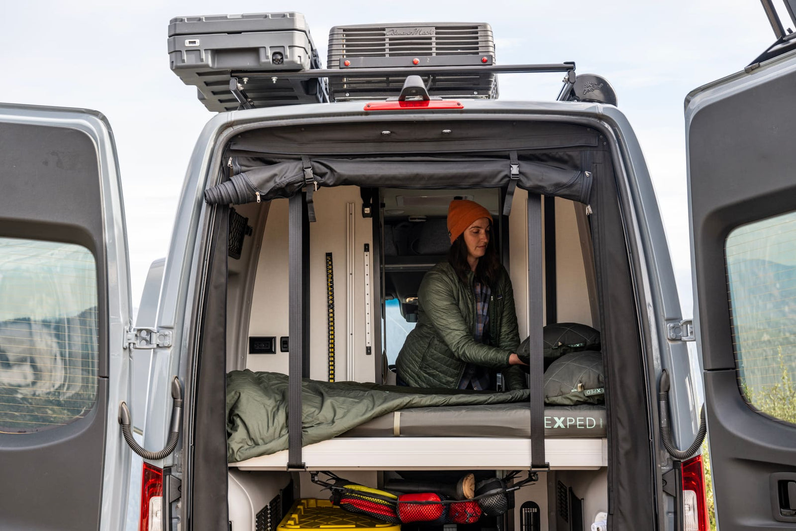 Eine Frau mit Mütze passt das Bettzeug und ihre EXPED Schlafmatte LuxeMat DUO M in einem Wohnmobil mit Regalen, Ausrüstung und Aufbewahrungsboxen an, bereit für ein Outdoor-Abenteuer, gesehen von den offenen Hintertüren.