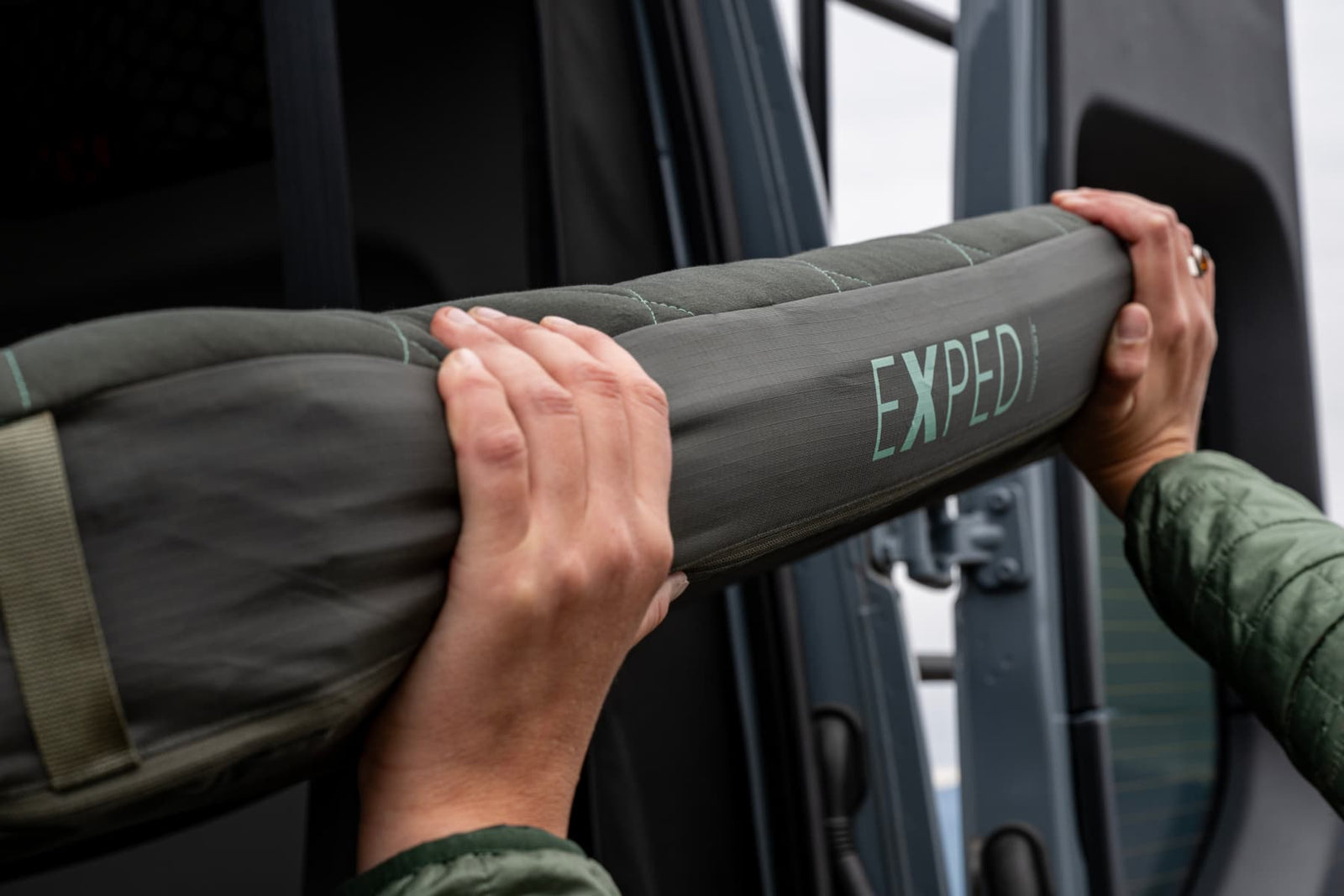 Nahaufnahme von zwei Händen, die die grüne EXPED Schlafmatte LuxeMat DUO M in der Nähe einer offenen Fahrzeugtür halten. Diese komfortable Schlafmatte von EXPED ist ideal für Outdoor-Abenteuer und eignet sich perfekt für Reisen.