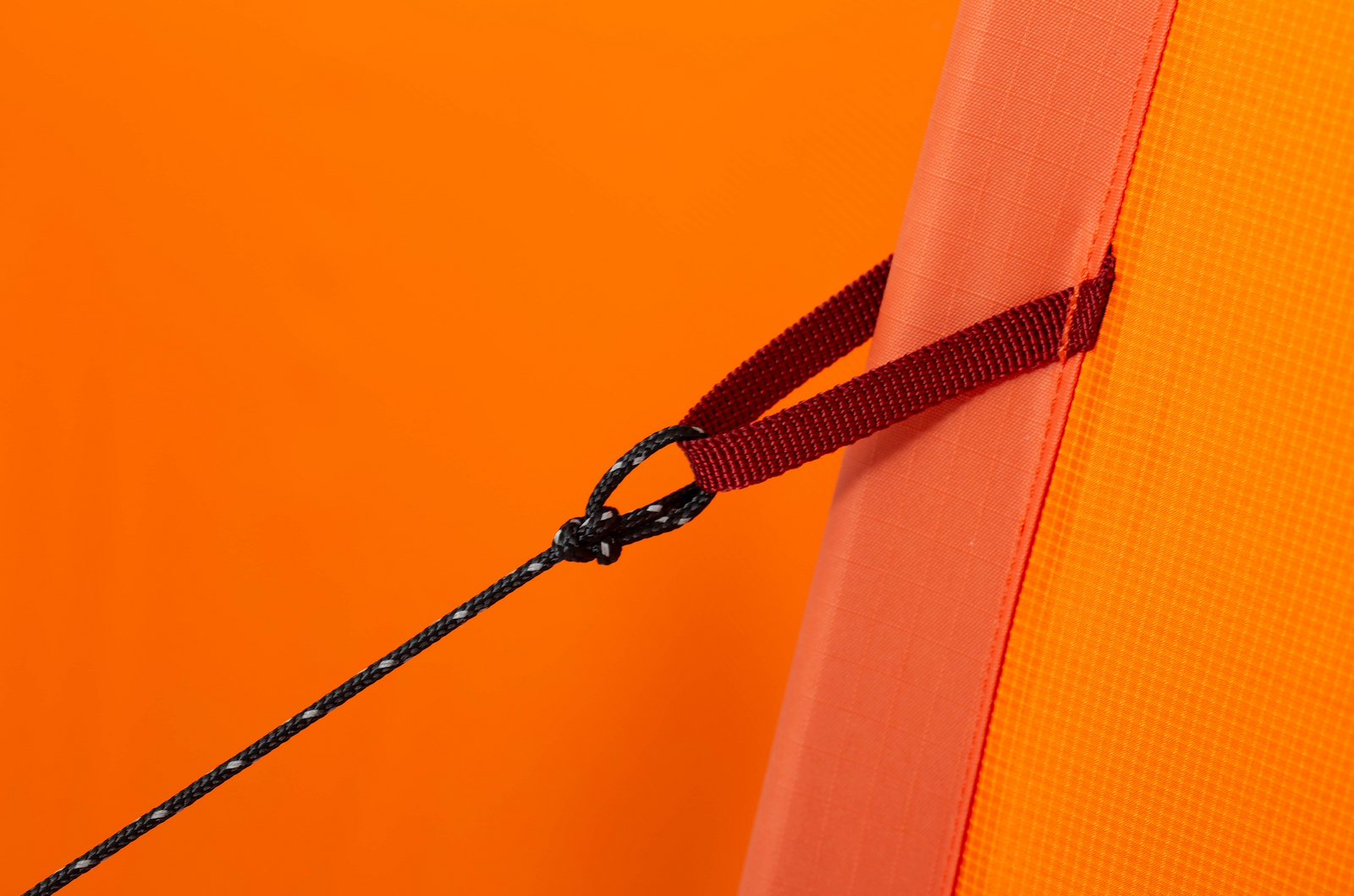 Nahaufnahme einer schwarzen Kordel, die mit einer roten Stoffschlaufe auf einer orangefarbenen, strukturierten Oberfläche verbunden ist - Teil des EXPED Zelt Polaris von EXPED - mit einem leuchtend orangefarbenen Hintergrund.