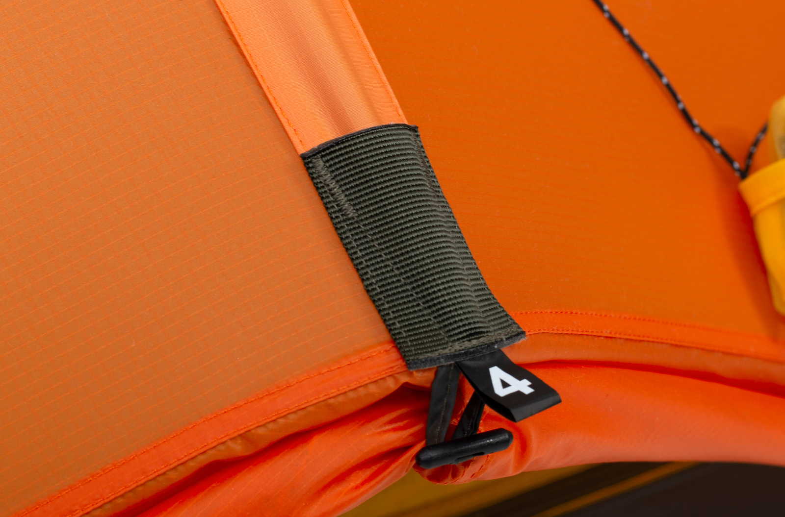 Nahaufnahme eines orangefarbenen Exped Zelt Polaris von EXPED, das ein schwarzes Band mit einem kleinen, an einer Naht befestigten Anhänger zeigt, wobei im Hintergrund ein Teil eines gelben Objekts und eine schwarze Kordel sichtbar sind.