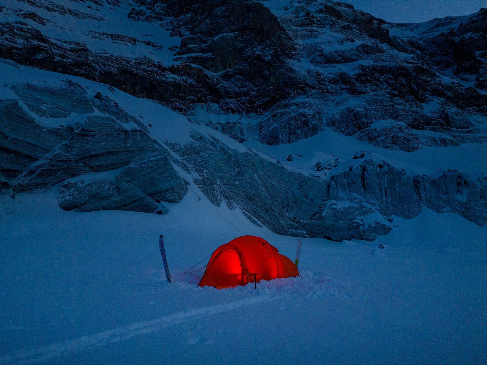 Ein EXPED Zelt Polaris leuchtet orange auf dem verschneiten Boden in der Dämmerung, umgeben von eisigen Felsen und schroffen, schneebedeckten Gipfeln. Die Skier stehen neben diesem 4-Jahreszeiten-Expeditionszelt von EXPED und fangen den Geist des abgelegenen Wintercampings ein.