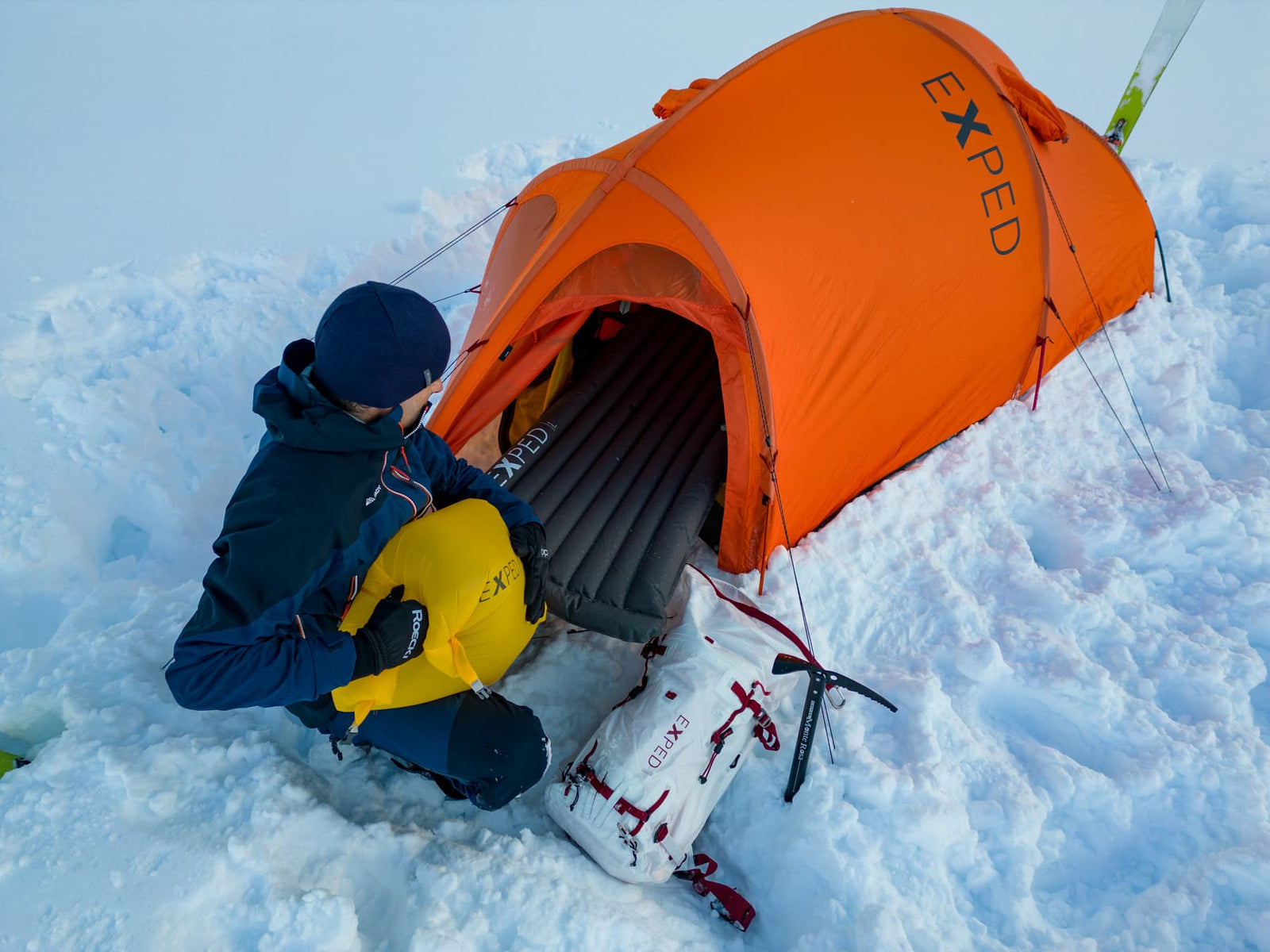 Eine Person in Winterausrüstung kniet im Schnee neben einem orangefarbenen Exped Zelt Polaris von EXPED und bläst eine Schlafmatte auf. Ein weißer Rucksack und ein Eispickel liegen in der Nähe auf dem Schnee.
