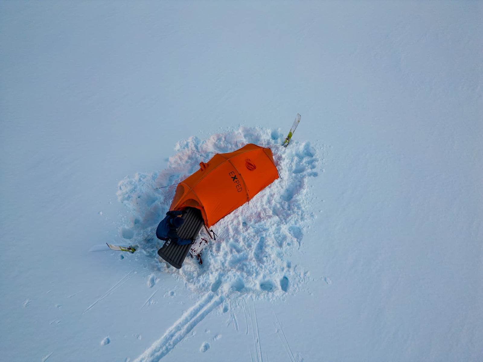 Ein EXPED Zelt Polaris in leuchtendem Orange steht auf unberührtem weißen Schnee, flankiert von zwei Paar Skiern und Ausrüstung, unter einem klaren Himmel.