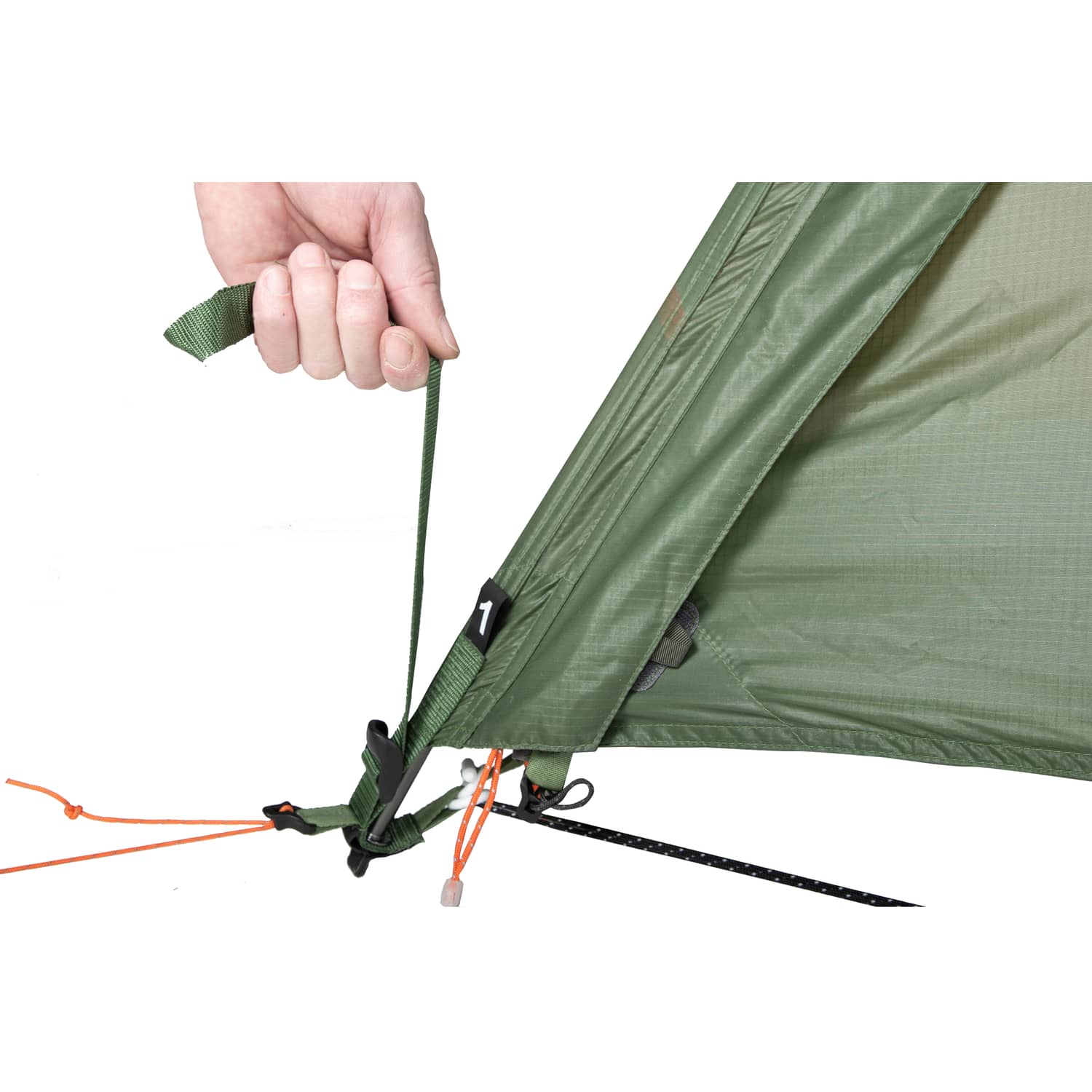 Nahaufnahme einer Hand, die an einem grünen Gurt zieht, um die Seite eines EXPED Venus III DLX Extreme-Zeltes mit orangefarbenen und schwarzen Abspannleinen am Boden zu befestigen.