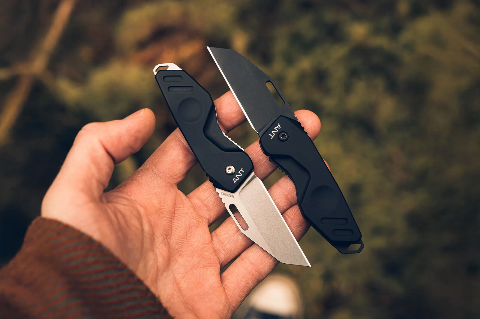 Eine Hand hält zwei kleine, schwarze Extrema Ratio Messer ANT Black M390 Stone Washed von EXTREMA RATIO, deren silberne M390 Stahlklingen sich vor einem verschwommenen Outdoor-Hintergrund aus Grün- und Brauntönen öffnen.