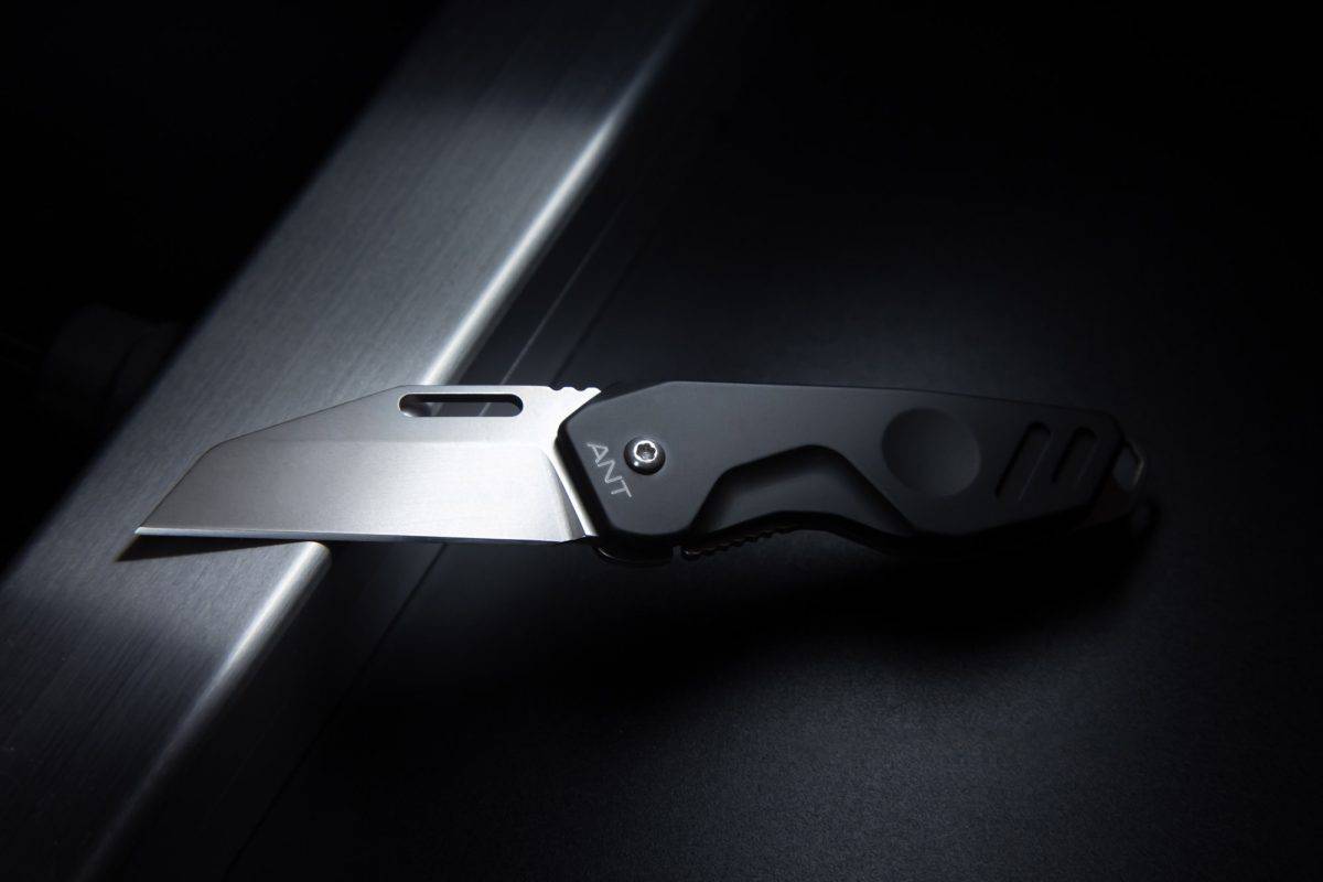 Das EXTREMA RATIO Messer ANT Black Stone Washed mit der schlanken Böhler N690 Stahlklinge und dem schwarzen Griff liegt teilweise offen auf einer dunklen Fläche, seine scharfe Kante und sein kantiges Design werden durch das Licht akzentuiert.