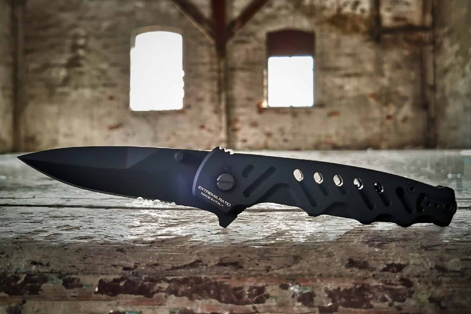 Ein schwarzes EXTREMA RATIO Extrema Ratio Messer Caimano Nero N.A. taktisches Klappmesser mit teilgezahnter Klinge ruht auf verwittertem Holz vor verschwommenen Bogenfenstern und zeigt die robuste Vielseitigkeit des Caimano Nero.