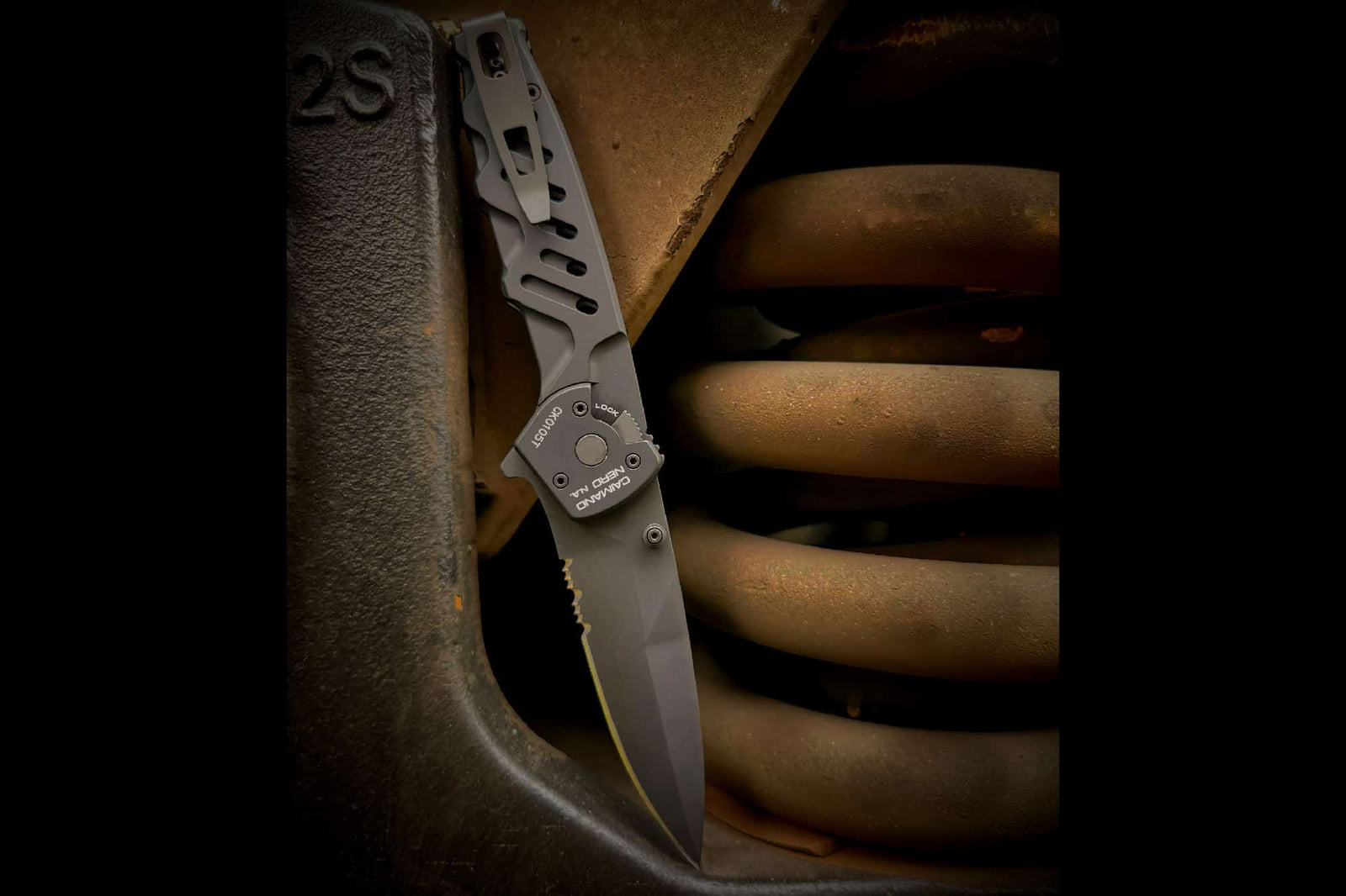 Das Extrema Ratio Messer Caimano Nero N.A. Klappmesser von EXTREMA RATIO, mit einer teilweise gezahnten schwarzen Klinge, steht auf einer großen, rostigen Spiralfeder und einer Metalloberfläche für einen robusten industriellen Look unter dramatischer Beleuchtung.