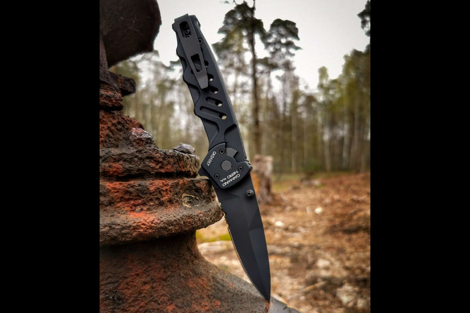 Ein EXTREMA RATIO Extrema Ratio Messer Caimano Nero N.A. mit teilgezahnter schwarzer Klinge ist im Freien in eine rostige Metalloberfläche eingebettet, Wald und erdiger Boden verschwimmen im Hintergrund - ein echtes taktisches Messer.