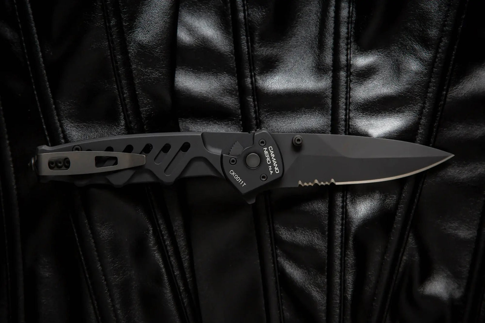 Das EXTREMA RATIO Extrema Ratio Messer Caimano Nero N.A., ein schwarzes taktisches Klappmesser mit teilgezahnter Klinge, wird auf einer glänzend schwarzen Lederoberfläche gezeigt.