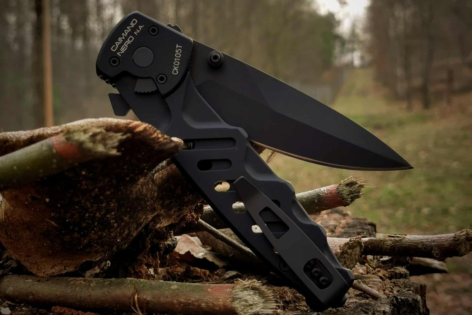 Das EXTREMA RATIO Extrema Ratio Messer Caimano Nero N.A., ein schwarzes taktisches Messer mit teilweise gezahnter Klinge, wird im Freien auf abgeschnittenen Ästen vor einem verschwommenen Waldhintergrund präsentiert.
