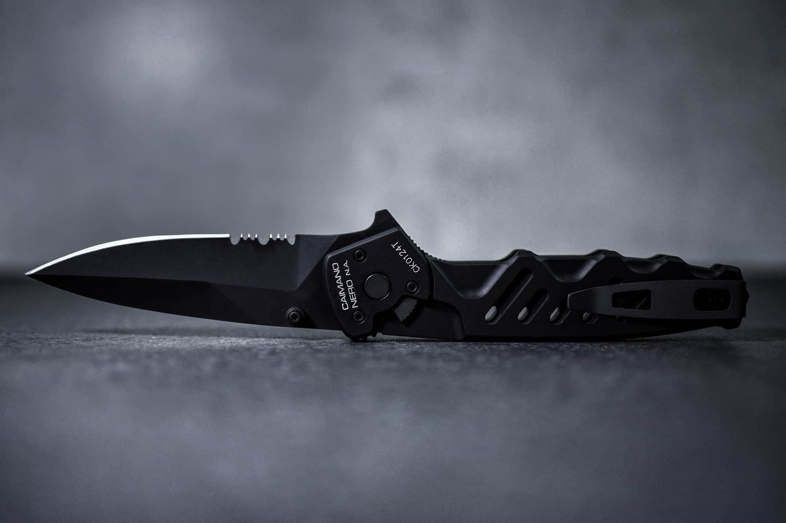 Ein Extrema Ratio Messer Caimano Nero N.A. von EXTREMA RATIO mit teilweise gezackter Klinge und strukturiertem Griff ruht auf einer glatten grauen Oberfläche vor einem verschwommenen grauen Hintergrund.