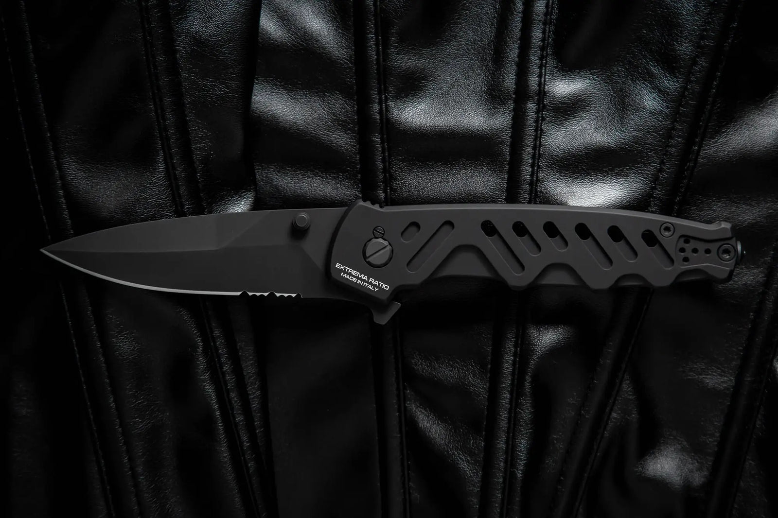 Das EXTREMA RATIO Extrema Ratio Messer Caimano Nero N.A., ein schwarzes Klappmesser mit teilgezahnter Klinge, liegt auf einer genähten schwarzen Lederoberfläche.